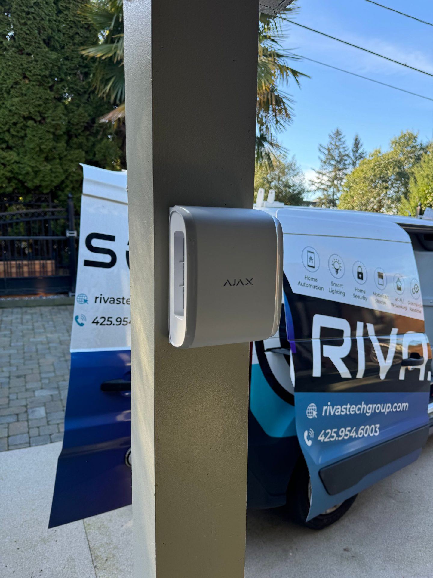 A white Ajax outdoor motion detector on a gray post with a Rivas Technology Group service van parked behind in sun