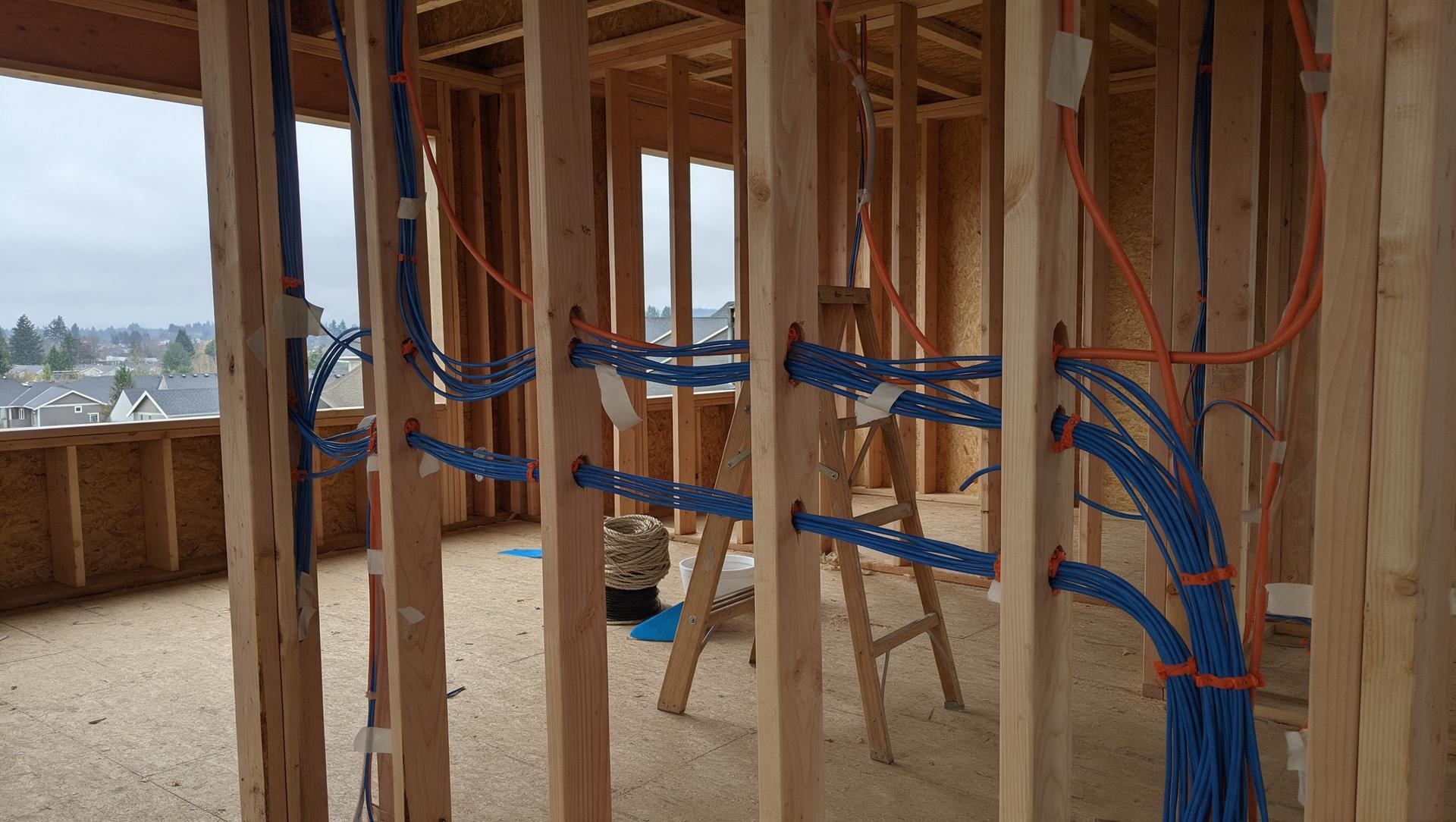 Bundles of blue low-voltage cable and orange conduit run through the wood studs of a house under construction