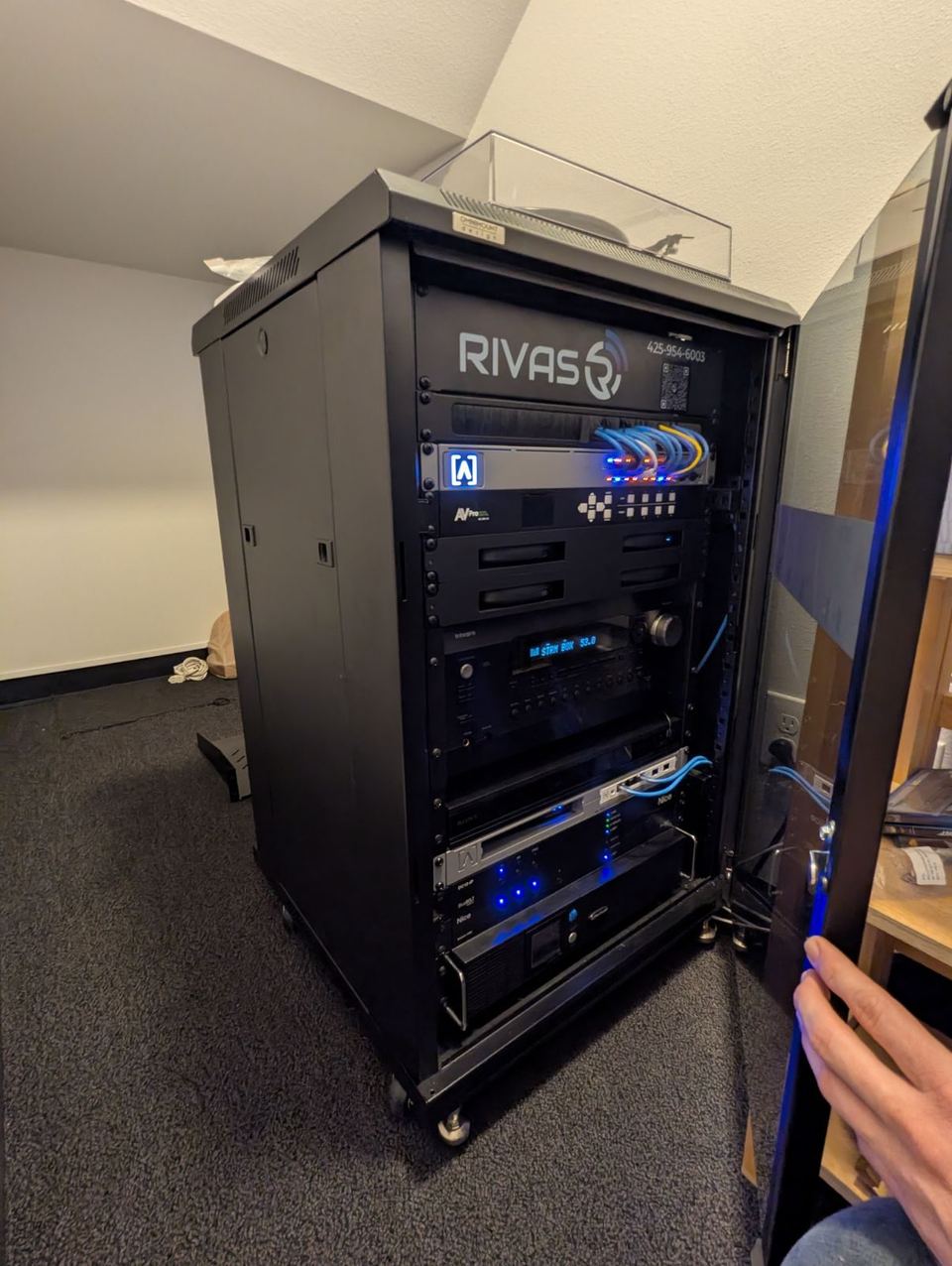 A black equipment rack with a Rivas Technology Group logo panel, blue-lit network gear, a receiver and patch cables in a carpeted room